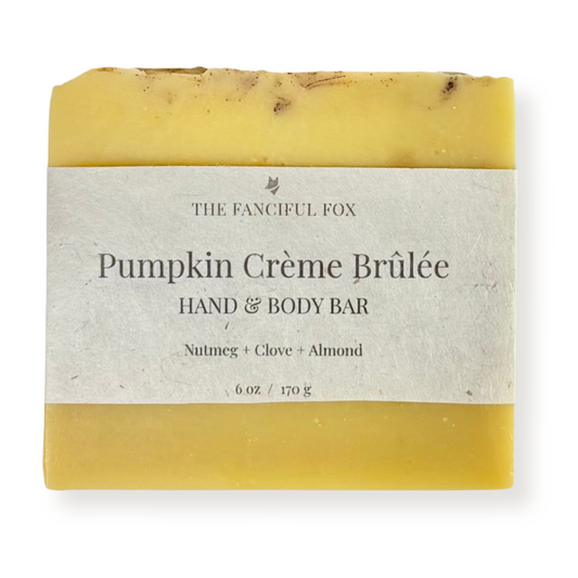 A rectangular hand and body bar of soap with a cream-colored paper label reading 'THE FANCIFUL FOX Pumpkin Crème Brûlée HAND & BODY BAR'. The soap has a warm yellow base with darker brown and black flecks throughout, set against a clean white background.
