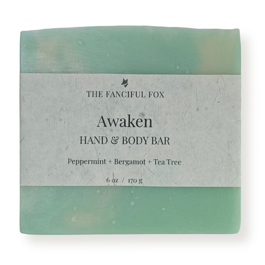 A rectangular hand and body bar wrapped in a light gray paper label that reads 'THE FANCIFUL FOX Awaken HAND & BODY BAR' with scent notes 'Peppermint + Bergamot + Tea Tree' and weight '6 oz / 170 g'. The soap itself has a soft mint green and pale yellow marbled pattern visible at the top and bottom edges.