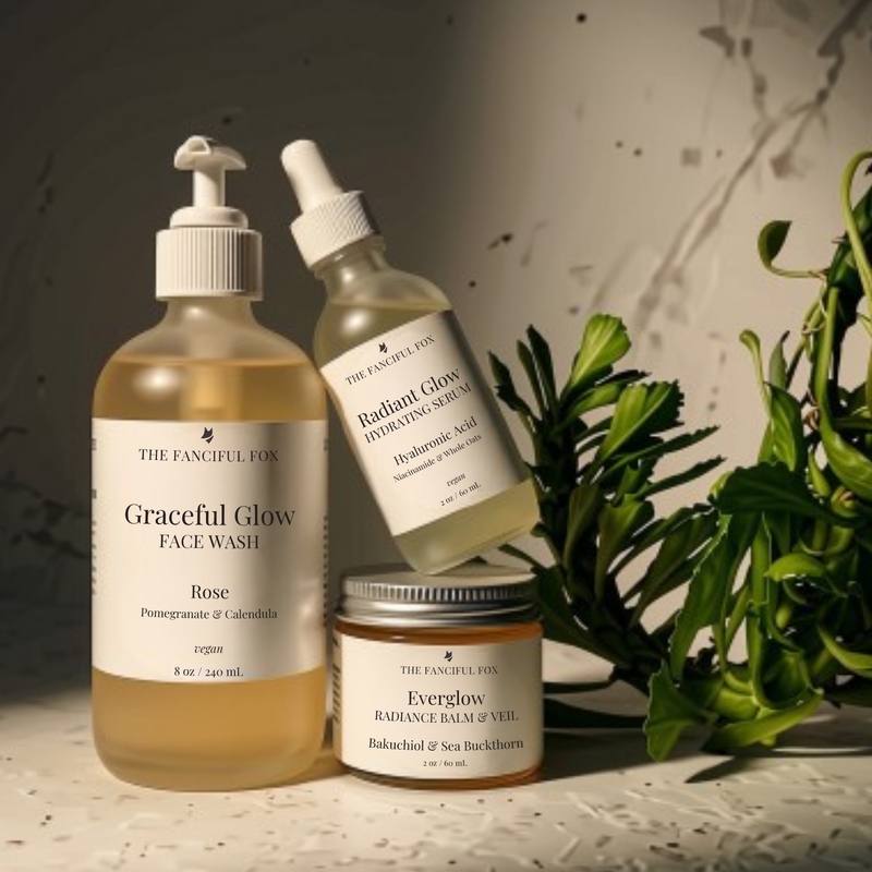 Three bottles of The Fanciful Fox skincare products arranged on a textured gray surface with green leaves in the foreground. The products include a large amber bottle of Golden Glow Facial Wash, a small jar of Everglow Radiance Balm, and a dropper bottle of Radiant Glow Serum.