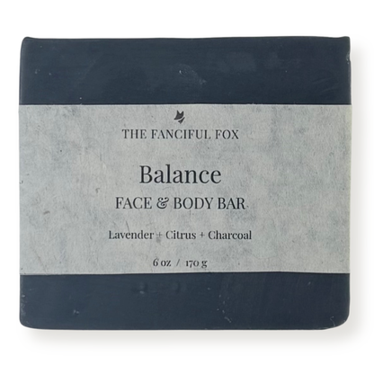 A rectangular bar of soap wrapped in a light gray paper label, with dark navy blue top and bottom bands. The label reads 'THE FANCIFUL FOX Balance FACE & BODY BAR' and 'Lavender + Citrus + Charcoal'.