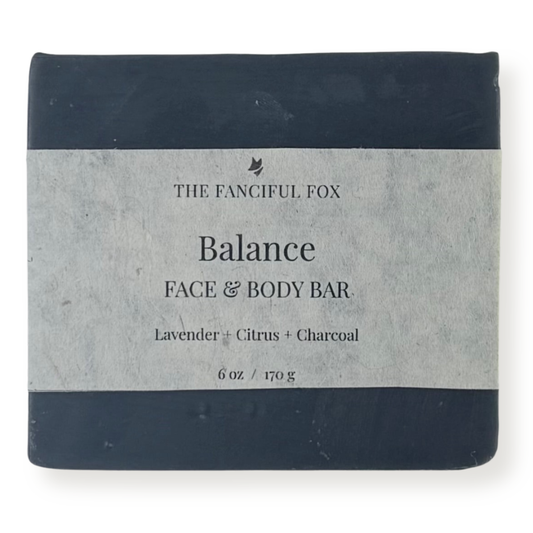 A rectangular bar of soap wrapped in a light gray paper label, with dark navy blue top and bottom bands. The label reads 'THE FANCIFUL FOX Balance FACE & BODY BAR' and 'Lavender + Citrus + Charcoal'.