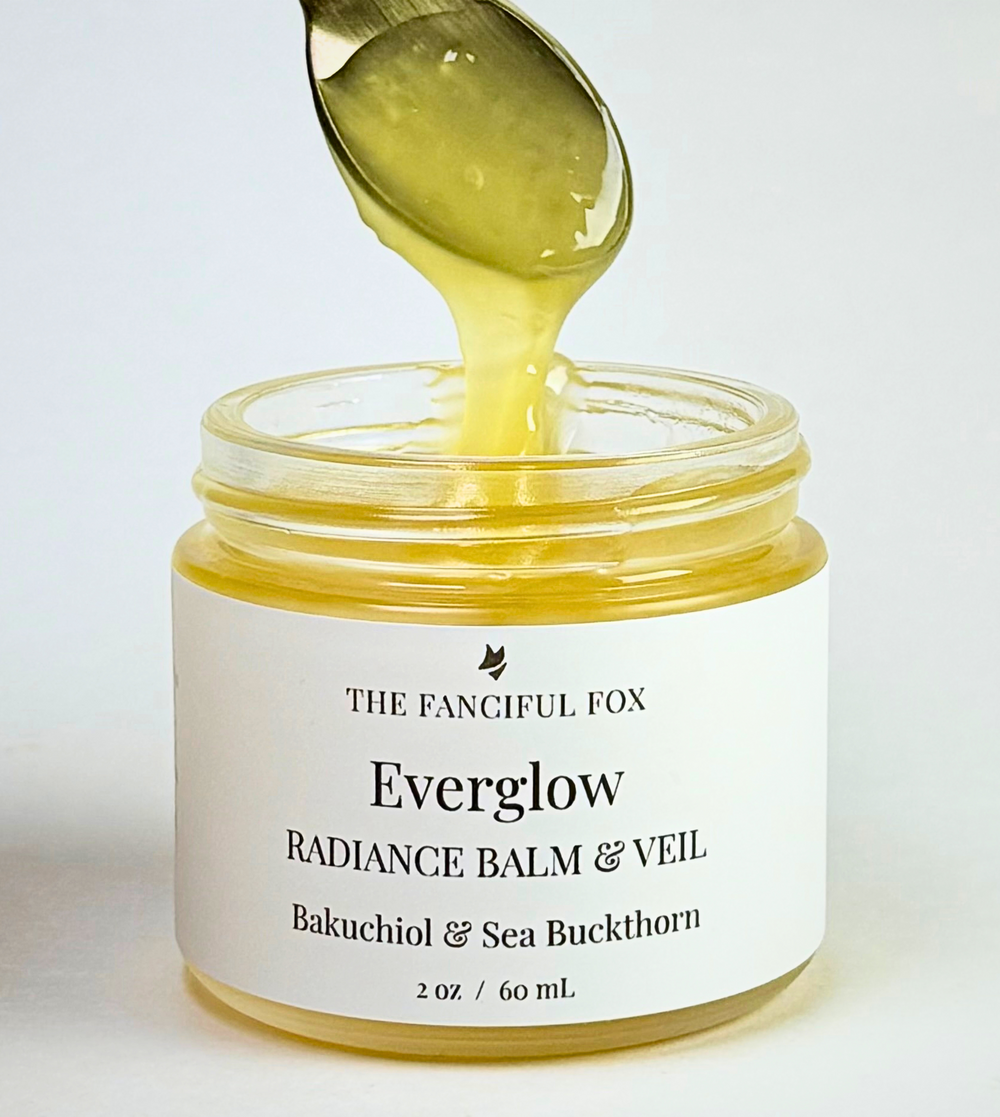 A glass jar of Everglow Radiance Balm & Veil by The Fangful Fox, showing a golden-yellow balm being drizzled from a spoon above the open jar, set against a clean white background.